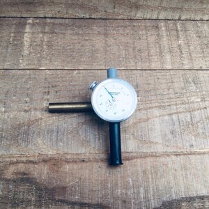 May include: A dial indicator with a white face, black numerals, and a blue needle. It features a black stem and a brass-coloured extension. This precision measuring tool is used in engineering and manufacturing.