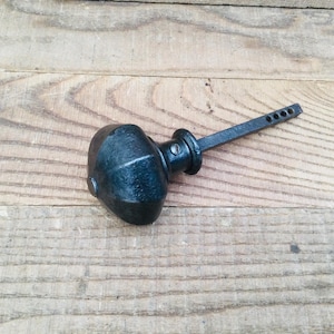 May include: A black, antique-style door knob with a rounded handle and a long, flat stem with several holes. The knob is made of metal and has a slightly textured surface. It rests on a wooden surface.