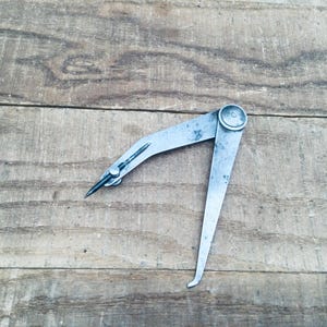 May include: A vintage metal caliper tool, featuring a pointed tip and curved leg. The tool is silver-toned and appears to be made of steel. It is resting on a weathered wooden surface, showcasing its age and use.