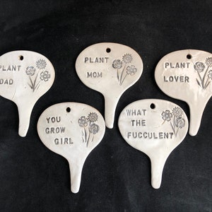 Puede incluir: Cinco marcadores de plantas de cerámica blanca con letras negras y diseños florales. Los marcadores dicen "Plant Dad", "Plant Mom", "Plant Lover", "You Grow Girl" y "What the Fuccolent".
