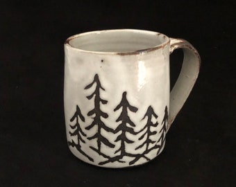 May include: White ceramic mug with a black silhouette of a forest of pine trees. The mug has a rounded body and a handle.