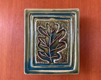 May include: Square ceramic tile with a raised oak leaf design. The tile features a dark green leaf with blue and white accents, set within a rectangular frame of green and blue. The tile is a decorative piece.