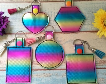 May include: Five different shaped keychains with rainbow holographic fabric. The keychains are a heart, a hexagon, a circle, a rectangle, and a square. Each keychain has a silver chain and a tassel.