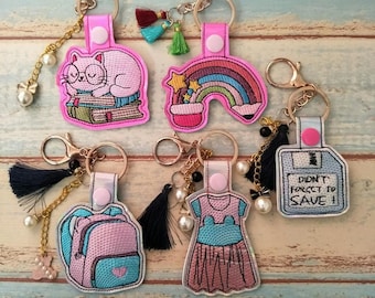 Puede incluir: Cinco llaveros rosas con diferentes diseños bordados. Los diseños incluyen un gato durmiendo sobre una pila de libros, un arcoíris, una mochila, un vestido y un disquete con el texto "¡No olvides guardar!"
