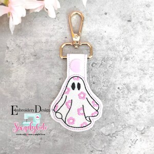 May include: A white ghost keychain with pink flowers embroidered on it. The ghost has two black eyes and a pink button on the top. The keychain is attached to a gold metal clip.