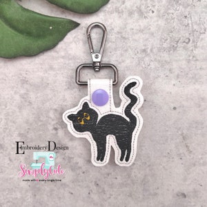 May include: Black cat keychain with a purple button and a silver clip. The keychain is made of felt and has a stitched design. The text "Simply Cute" is embroidered on the keychain.