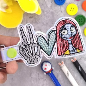 May include: A white iridescent patch with an embroidered peace sign, heart, and a character from the movie Nightmare Before Christmas. The peace sign is made of a skeleton hand, the heart is teal, and the character is Sally with red hair and a yellow dress.