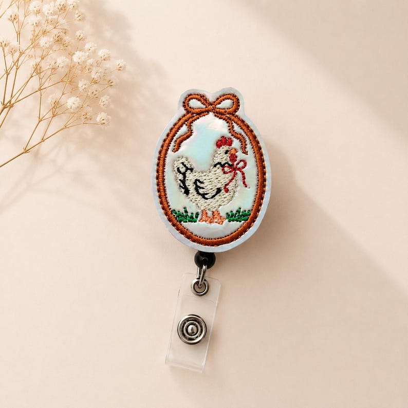 Op de afbeelding: Een ovale badgehouder met een geborduurde witte kip met een rode kam en een klein rood strikje om de nek. De kip is omlijst door een bruine geborduurde rand met een strik aan de bovenkant, op een lichtblauwe achtergrond met groen gras.