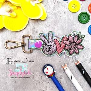 May include: A keychain with a peace sign, heart, and flower design. The word "love" is incorporated into the design. The keychain has a gold-colored clasp and a purple button. The text "Embroidery Design" and "Simply Cute" are also visible.