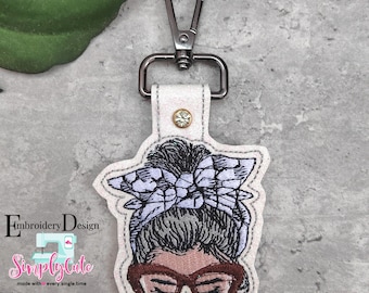 May include: A white key chain with a colorful embroidered design of a woman with a messy bun and glasses. The design includes a blue and white checkered bow.