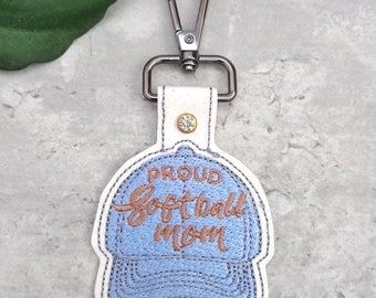 May include: A white keychain with a blue embroidered baseball cap that says "Proud Softball Mom".