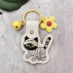 May include: A Halloween-themed keychain featuring an embroidered mummy cat design. The keychain includes a yellow bell, a yellow flower charm with a red center, and a cream-colored tag with a decorative gem. The cat has black ears and a striped tail.