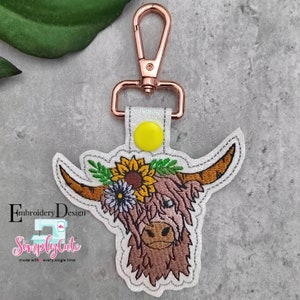 May include: A brown and white Highland cow head with a sunflower and flower crown, embroidered on a white glittery fabric keychain with a rose gold metal clasp.