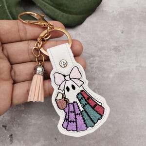 Peut inclure: Un porte-cl&eacute;s en cuir blanc avec un fermoir dor&eacute; et un pompon p&ecirc;che. Le porte-cl&eacute;s pr&eacute;sente un motif de fant&ocirc;me brod&eacute; avec un n&oelig;ud rose, tenant une tasse. La robe patchwork du fant&ocirc;me est violette, verte et rouge.