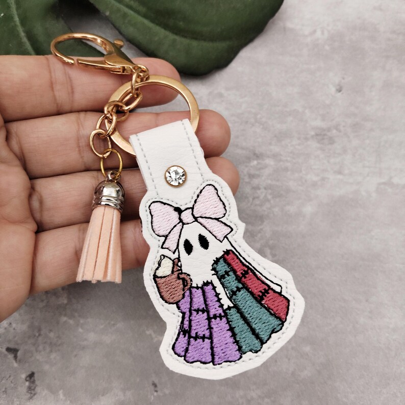 Peut inclure: Un porte-cl&eacute;s en cuir blanc avec un fermoir dor&eacute; et un pompon p&ecirc;che. Le porte-cl&eacute;s pr&eacute;sente un motif de fant&ocirc;me brod&eacute; avec un n&oelig;ud rose, tenant une tasse. La robe patchwork du fant&ocirc;me est violette, verte et rouge.