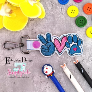 Op de afbeelding: Een witte stoffen sleutelhanger met een zilveren sluiting, waarop een blauw vredesteken, een roze hart en een blauwe pootafdruk met een klein roze hartje erin geborduurd zijn. De tekst "Simply Cute" is onder het ontwerp geborduurd.