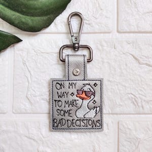 以下が含まれることがあります： シルバーの金属製のリングと、サングラスをかけた白いアヒルが刺繍されたグレーのファブリックキーホルダー。キーホルダーには「On my way to make some bad decisions」というテキストが刺繍されています。