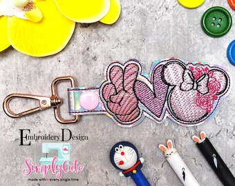 Puede incluir: Un llavero bordado de color rosa y blanco con la palabra "LOVE" en un diseño de signo de paz, corazón y orejas de ratón. El llavero está unido a un cierre de metal plateado y dorado.