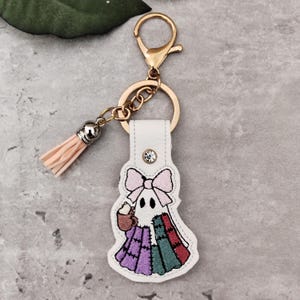 以下が含まれることがあります： ゴールドトーンの留め金とリングが付いた、白い革のキーホルダー。キーホルダーは、ピンクのリボンが付いた刺繍された幽霊のデザインで、コーヒーカップを持ち、パッチワークのキルトを着ています。 桃色のタッセルがチェーンからぶら下がっています。