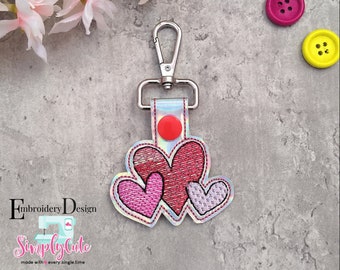 Puede incluir: Un llavero plateado con un diseño de corazón bordado en rosa, rojo y morado. Los corazones están superpuestos. El llavero está unido a un anillo plateado.