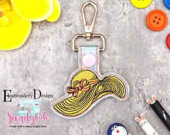 May include: A gold key chain with a yellow and orange embroidered sun hat charm. The charm is attached to a clear plastic keychain with a gold metal clasp. The text "Simply Cute" and "Embroidery Design" is visible on the image.