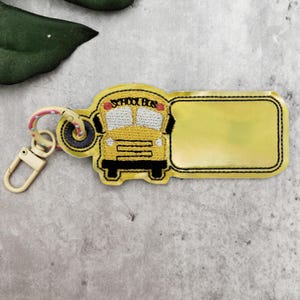 May include: Yellow school bus keychain with the words "SCHOOL BUS" embroidered on the top. The keychain has a blank yellow rectangle for personalization. The keychain is attached to a clasp and a ring.