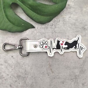 May include: White leather keychain with a black embroidered design of a dog and cat with a heartbeat line and paw print. The keychain has a silver metal clasp.