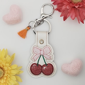 May include: A white leather keychain with a cherry design. The cherry design features a pink bow and two red cherries. The keychain has a silver clasp and a small orange tassel. A small rhinestone is set in the leather.
