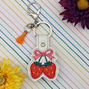 Può includere: Un portachiavi in pelle bianca con chiusura argentata e nappa arancione. Il portachiavi presenta un design a fragole rosse con un fiocco rosa e un piccolo strass. Le fragole hanno steli verdi.