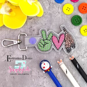 May include: A gray key chain with a peace sign, heart, and feather design. The key chain has a silver clip and a purple button. The text "Simply Cute" is visible in the image.