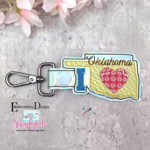 Puede incluir: Un llavero azul claro con un clip plateado. El llavero presenta un diseño bordado del estado de Oklahoma con las palabras "I love Oklahoma" y un corazón rojo con copos de nieve blancos.