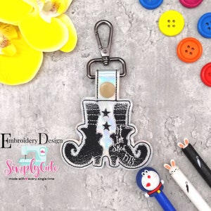May include: Black and white embroidered witch boot keychain with the text "If the shoe fits". The keychain is attached to a silver metal key ring.