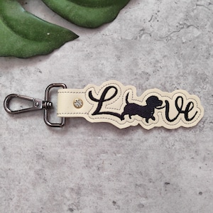 May include: A white leather keychain with a black embroidered dachshund and the word "Love". The keychain has a silver metal clasp.