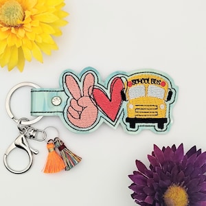 Peut inclure: Un porte-clés bleu et blanc tie-dye avec un signe de paix, un cœur rouge et un bus scolaire jaune avec le texte "School Bus". Le porte-clés a un anneau de clé argenté et deux glands orange et multicolores.
