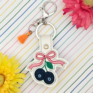 May include: A white leather keyring with a silver clasp and orange tassel. The keyring features a stitched design of two blueberries with a pink bow and green leaf. A small rhinestone is set on the white leather.