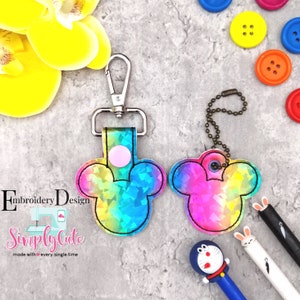 Peut inclure: Un porte-clés en forme de Mickey Mouse aux couleurs arc-en-ciel avec un fermoir argenté et une chaîne argentée. Le porte-clés est en tissu et a une finition holographique. Le texte "Simply Cute" est visible sur l'image.