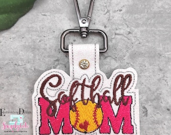 May include: White fabric keychain with a brown and pink embroidered design that says "Softball Mom" with a yellow softball in the middle. The keychain has a silver metal clasp.