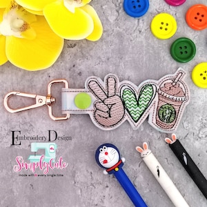 May include: A pink, green, and white embroidered keychain with a peace sign, heart, and a cup of coffee. The keychain has a gold metal clasp and is attached to a light blue and white fabric strap.