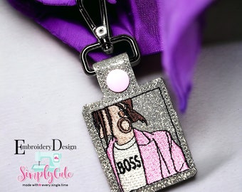 Puede incluir: Llavero de purpurina plateada con una imagen bordada en rosa y blanco de una persona que lleva una chaqueta rosa y una camiseta blanca con la palabra "BOSS" en ella.