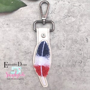 Puede incluir: Un llavero color crema con un cierre metálico y un diseño de pluma decorativa. La pluma está bordada con hilos azules, blancos y rojos. También se ve el texto "Embroidery Design" y "Simply Cute".