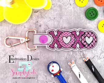 Puede incluir: Un llavero bordado en rosa y blanco con la palabra "XOXO" y dos corazones. El llavero está unido a un anillo de llave de metal dorado.