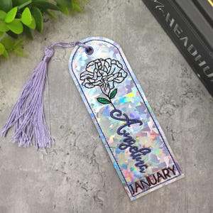 Könnte beinhalten: Ein silberner, schimmernder Lesezeichen mit einer weißen gestickten Blume und dem Namen "Angelina" sowie dem Monat "JANUARY", der in blauer Farbe gestickt ist. Das Lesezeichen hat einen lilafarbenen Quasten.