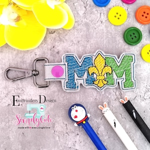 May include: A blue, green, and yellow embroidered keychain with the word "MOM" and a fleur-de-lis design. The keychain has a silver metal clasp.
