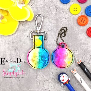 May include: Two rainbow iridescent keychains with a silver metal ring and a silver metal clasp. The keychain on the left has a yellow button. The keychain on the right has a silver metal ring. The keychains are on a gray background.