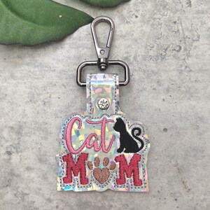 May include: A silver key chain with a sparkly iridescent charm that says "Cat Mom" in pink lettering with a black cat silhouette and a brown paw print.