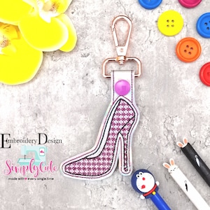 May include: A pink high heel shoe keychain with a gold metal clasp. The keychain is made of fabric and has a checkered pattern. The keychain is attached to a white fabric strap with a pink button.