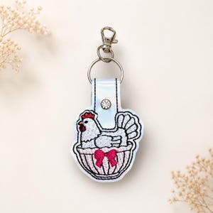 May include: A white iridescent keychain featuring an embroidered design of a white chicken sitting in a pink basket with a bright pink bow. The keychain includes a silver-toned metal lobster clasp and a circular ring, accented by a small clear rhinestone.