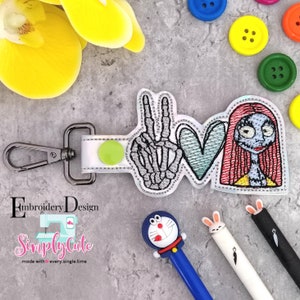 May include: A white keychain with a green circle and a silver clasp. The keychain features an embroidered design of a skeleton hand making a peace sign, a blue heart, and a cartoon character with red hair. The text "Simply Cute" is embroidered on the keychain.