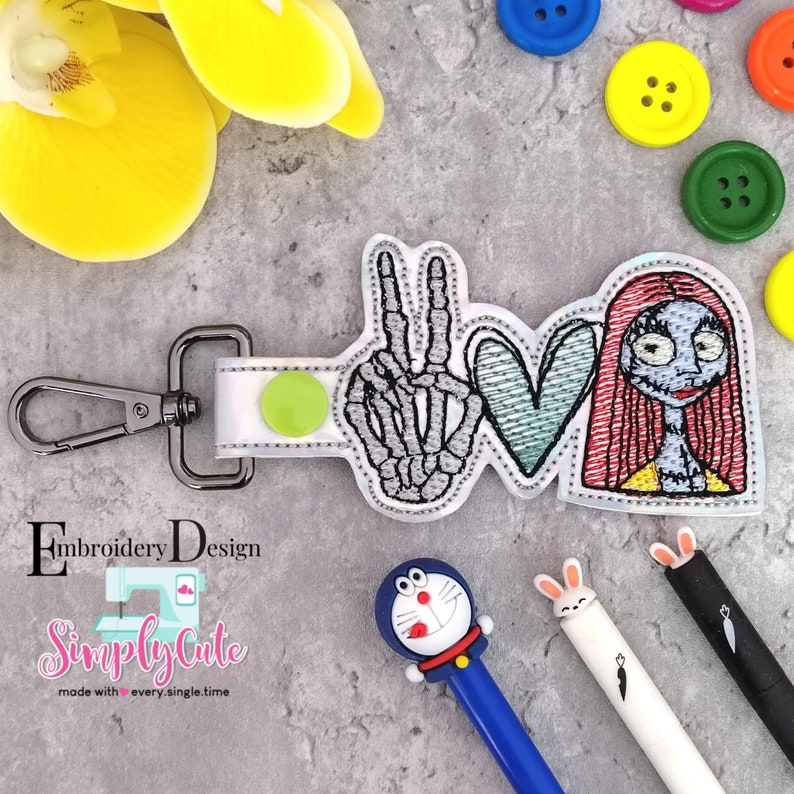 May include: A white keychain with a green circle and a silver clasp. The keychain features an embroidered design of a skeleton hand making a peace sign, a blue heart, and a cartoon character with red hair. The text "Simply Cute" is embroidered on the keychain.