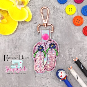 May include: A pink and white embroidered flip flop keychain with a green strap and blue flower embellishments. The keychain is attached to a silver metal key ring.
